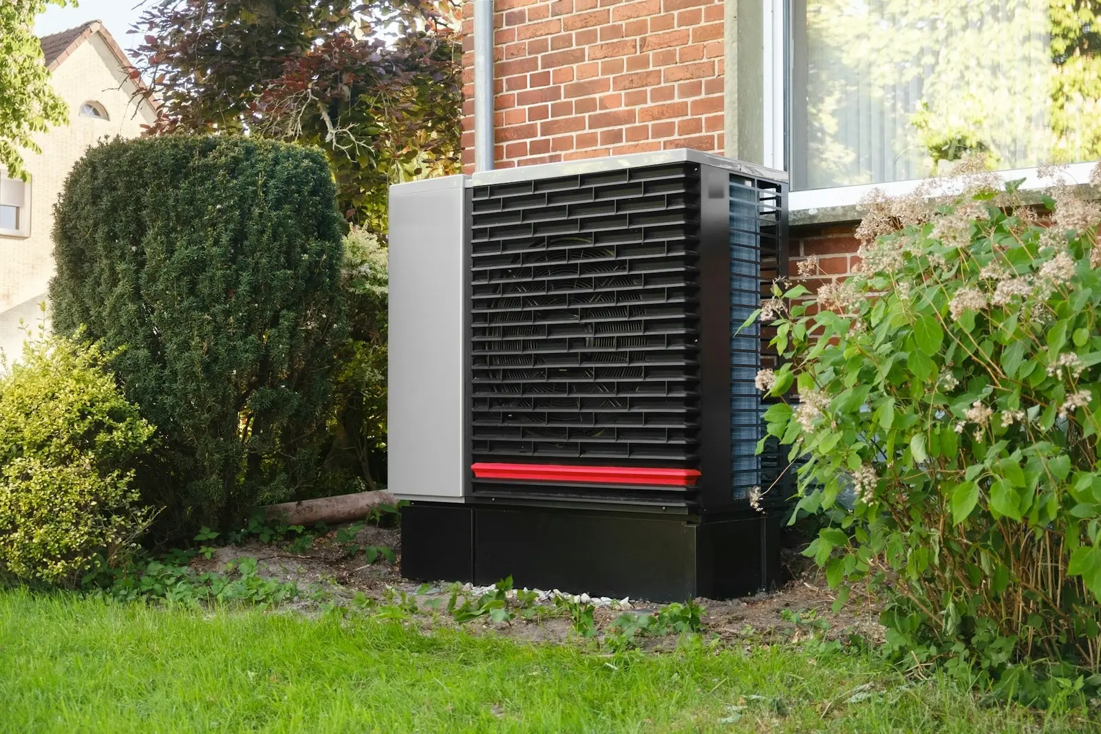 Een grijze warmtepomp met een zwart rooster in de tuin, tegen de bakstenen muur van een huis, tussen de struiken